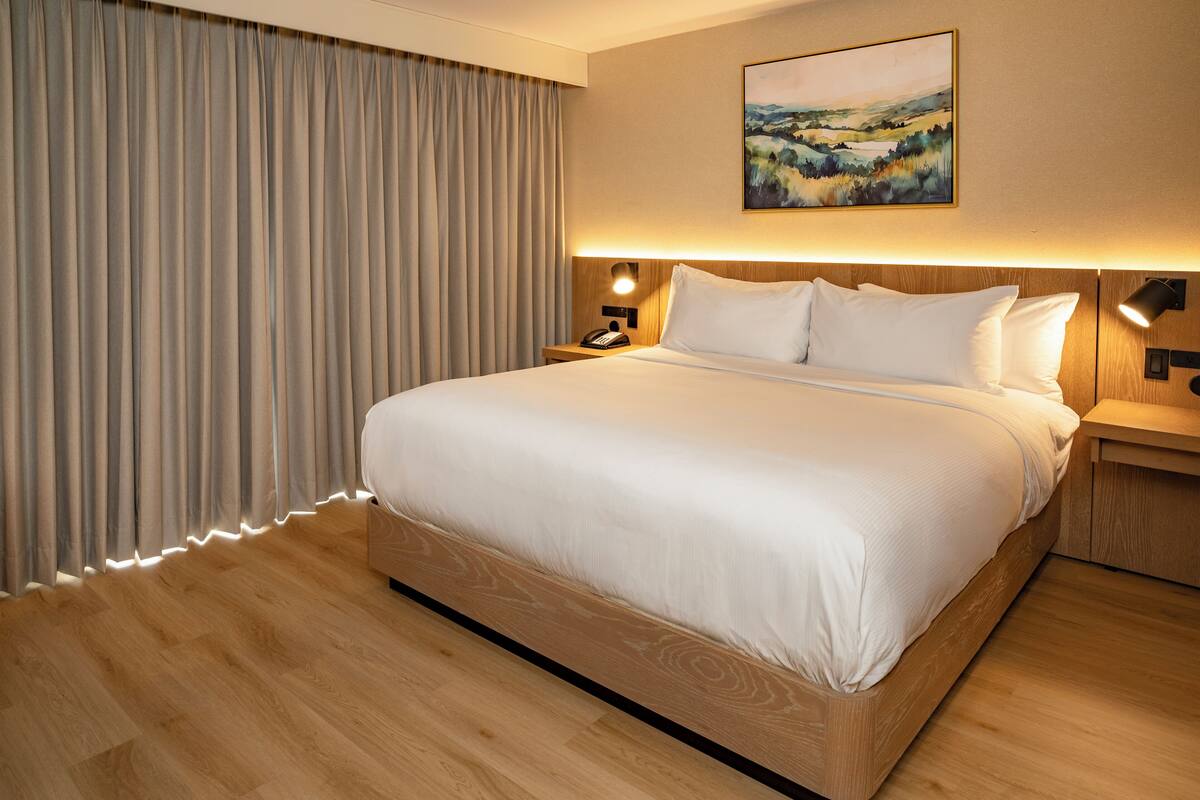 A hotel room with a large bed, wooden flooring, a nightstand with a lamp, and a painting on the wall.