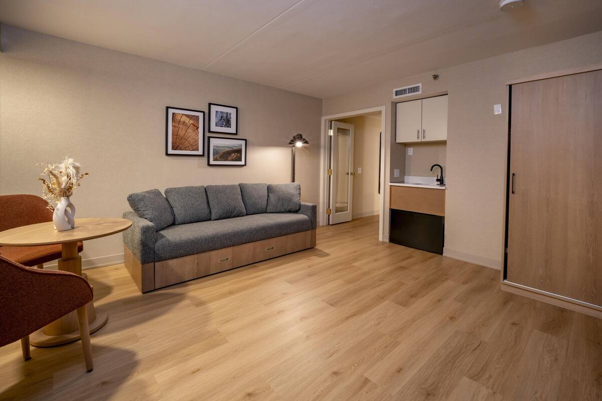 A modern living room with a grey sofa, a round wooden table, and a red chair. There are framed pictures on the wall and a lamp on the table.
