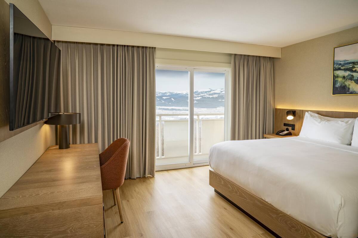 A hotel room with a large bed, a desk, a chair, a nightstand with a lamp, and a view of a snowy landscape through the window.