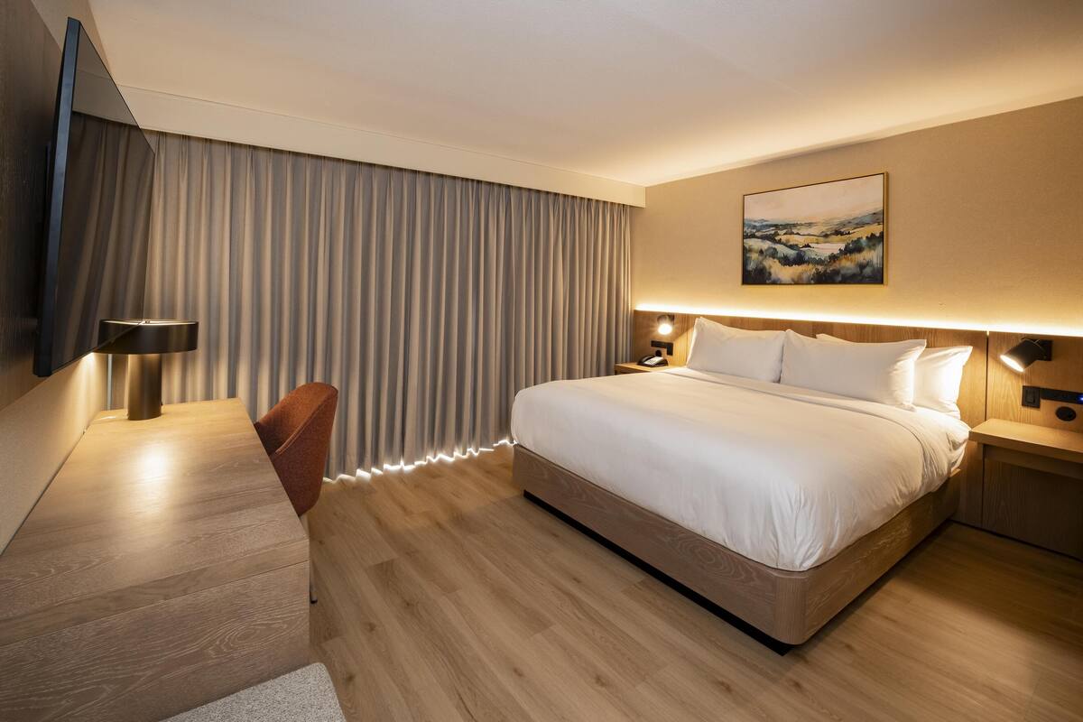 A modern hotel room with a large bed, a desk with a chair, a flat-screen TV mounted on the wall, and a painting above the bed.