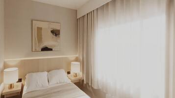 A hotel room with a large bed, two bedside tables with lamps, a painting on the wall, and a large window with curtains.