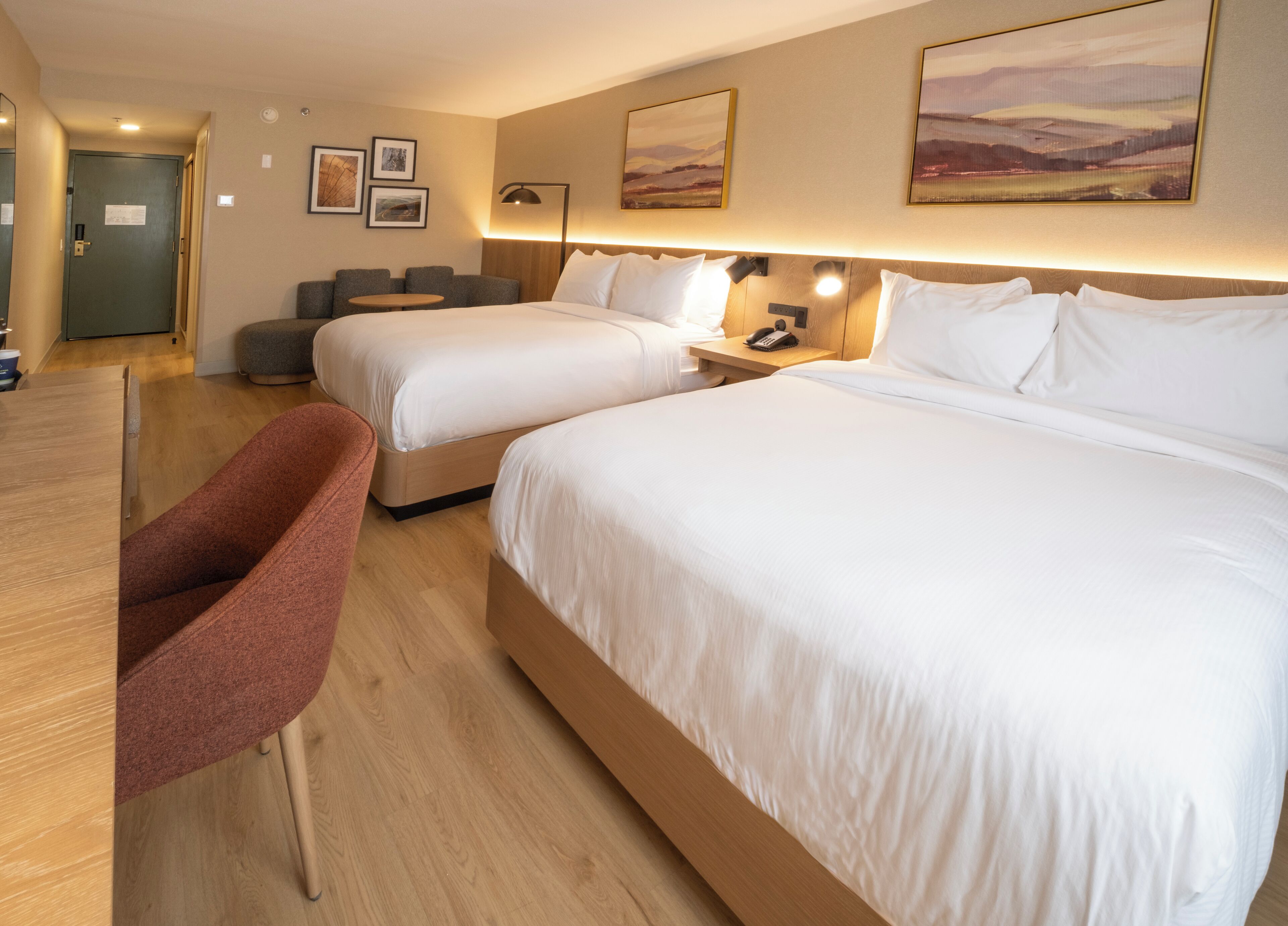 A hotel room with two beds, a desk, and a chair. There are framed pictures on the wall and a lamp on the bedside table.