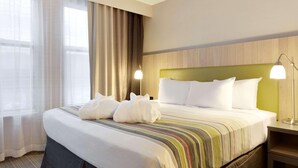 A hotel room with a large bed, a green headboard, two bedside lamps, and a window with sheer curtains.