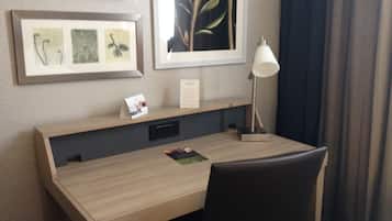 A hotel room with a wooden desk, a black chair, a desk lamp, and framed artwork on the wall.