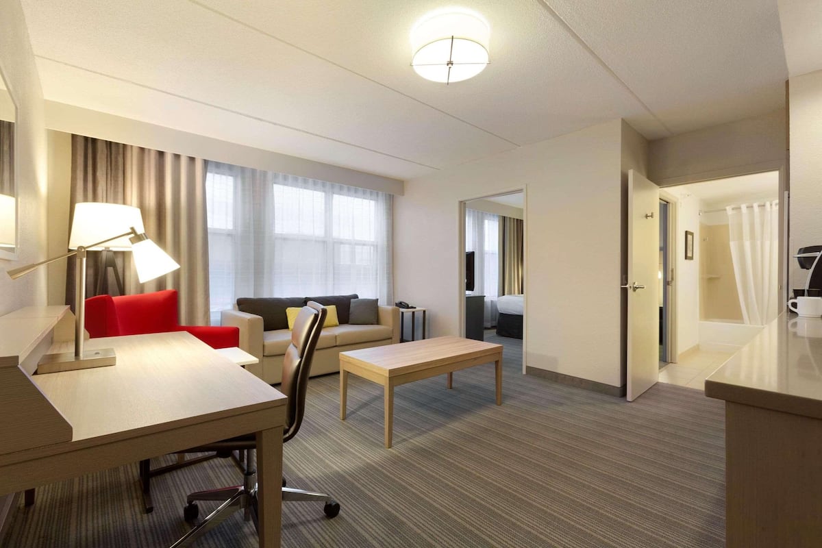 Hotellihuoneessa on työpöytä, tuoli, sohva ja pieni pöytä. Huoneessa on ikkuna, jossa on verhot, ovi, joka johtaa toiseen huoneeseen, ja kattovalaisin.