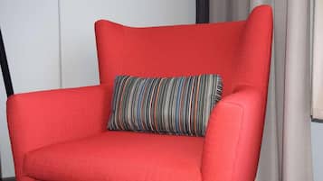 Un sillón rojo con un cojín a rayas frente a una pared blanca y una ventana con cortinas.