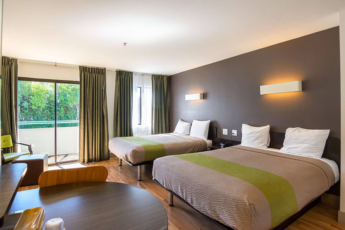 A hotel room with two beds, a dining area, and a balcony with curtains.