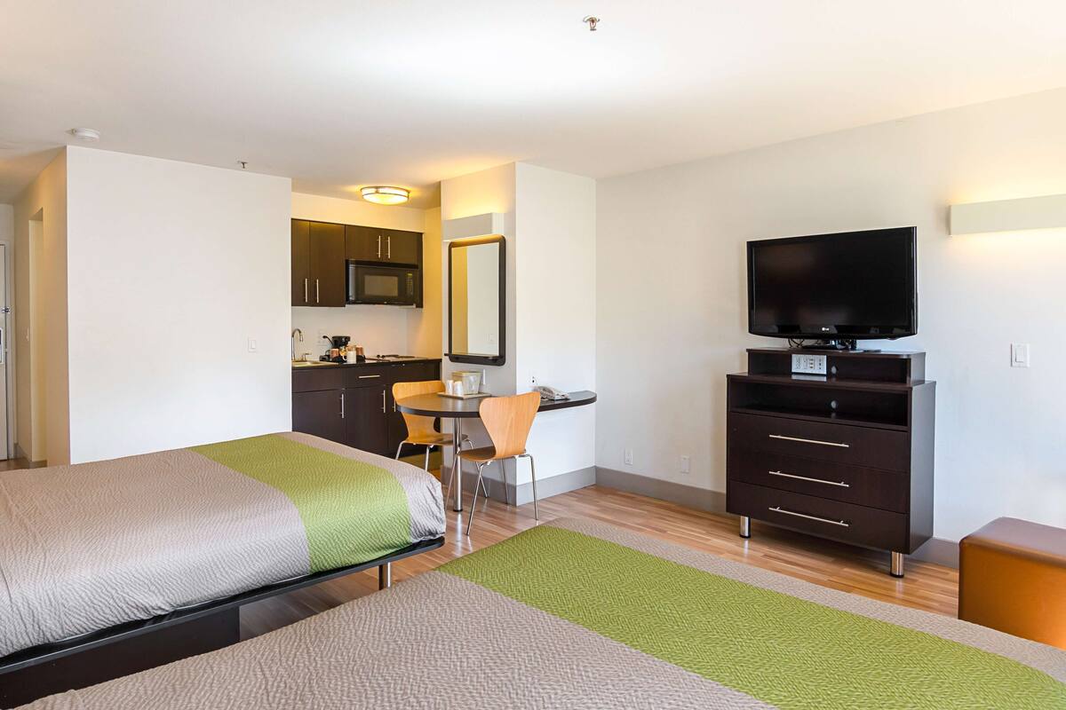A hotel room with two beds, a TV, a desk, and a kitchenette.