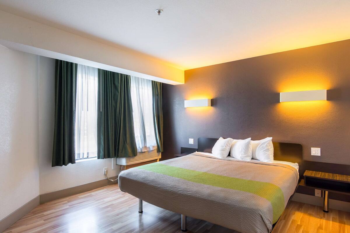 A hotel room with a large bed, two bedside tables, a nightstand, a window with curtains, and wall-mounted lighting.