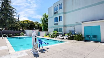 Una piscina amb una cadira de socorrista i una escala de piscina, envoltada de gandules i un edifici amb una porta blava.