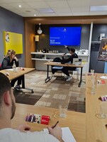 Uma sala de conferências com uma tela grande exibindo uma apresentação, mesas com copos d'água e uma pessoa apresentando.