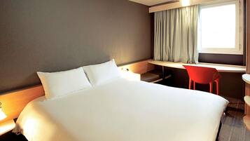 A hotel room with a large bed, a desk, a chair, and a window with curtains.