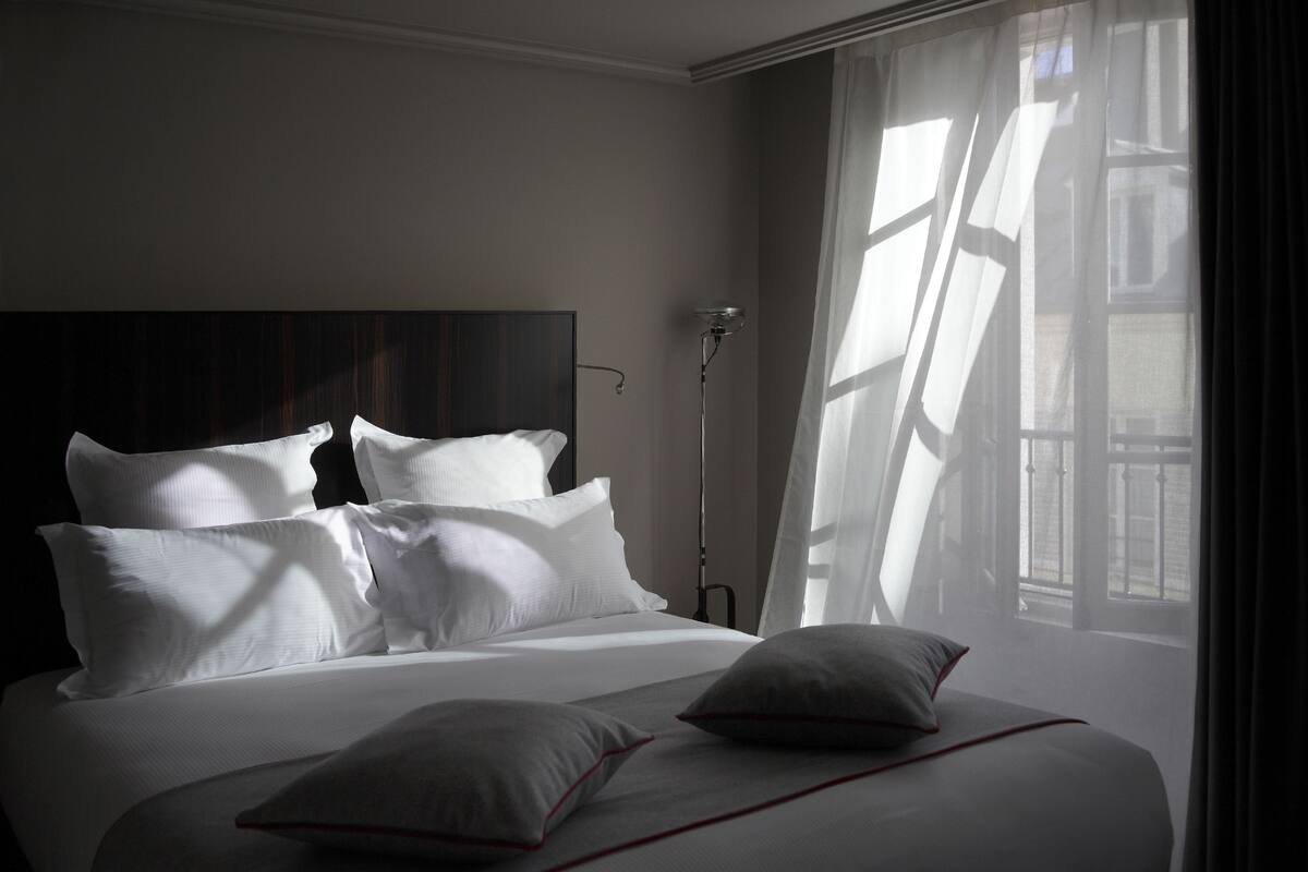 A neatly made bed with white linens and pillows, a bedside lamp, and a window with sheer curtains allowing natural light.