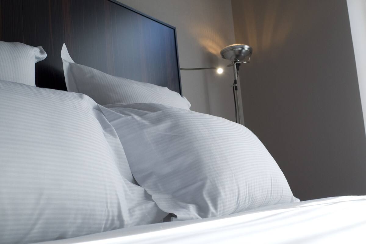 A neatly made bed with white linens and pillows, a dark headboard, and a floor lamp in a modern hotel room.