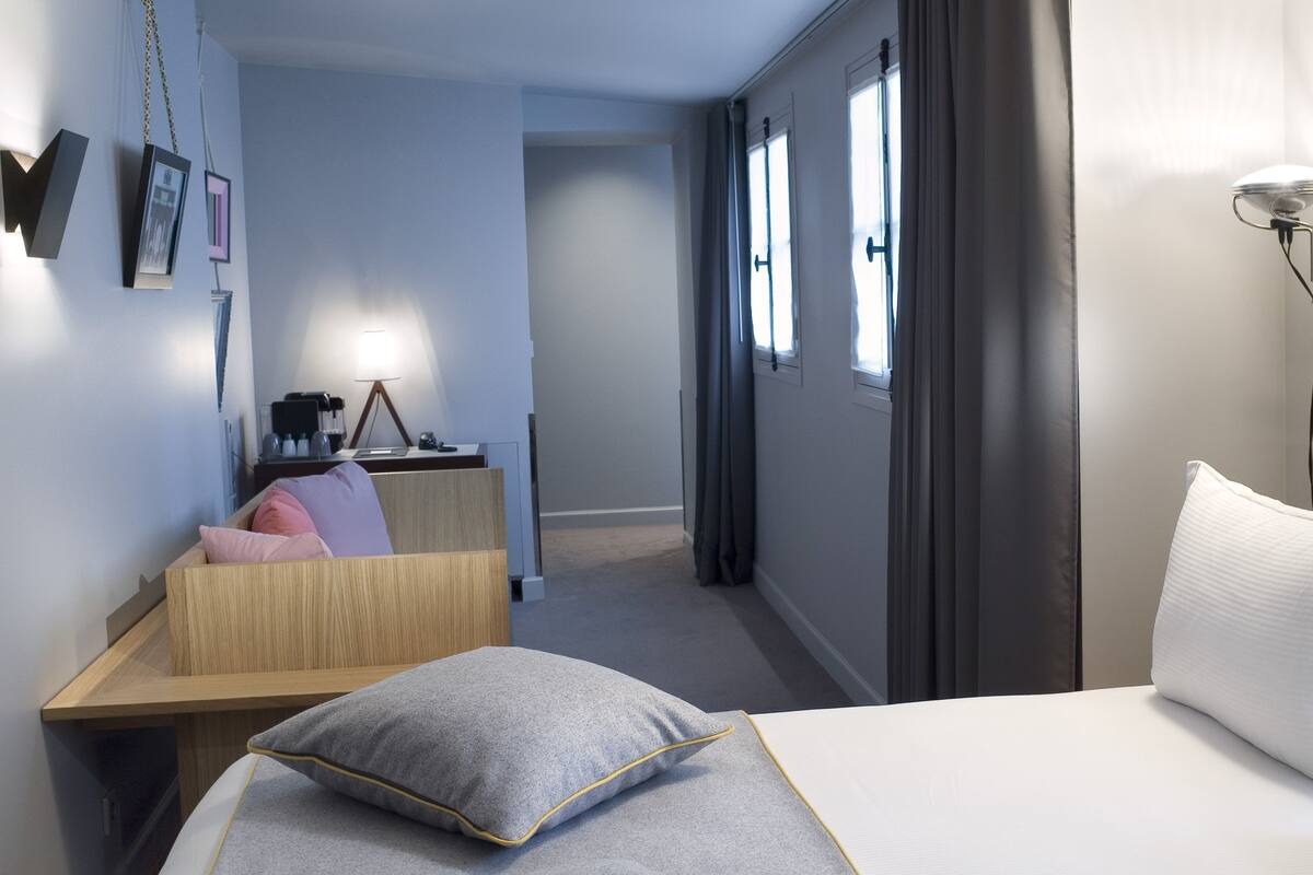 A hotel room with a bed, bedside table, lamp, and a view of the hallway.