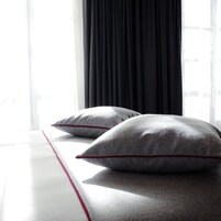 A bed with two pillows and a red accent on the bedspread, positioned next to a window with black curtains.