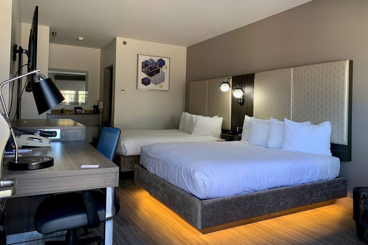 A hotel room with a desk, two beds, a chair, a desk lamp, a wall clock, and a framed picture.