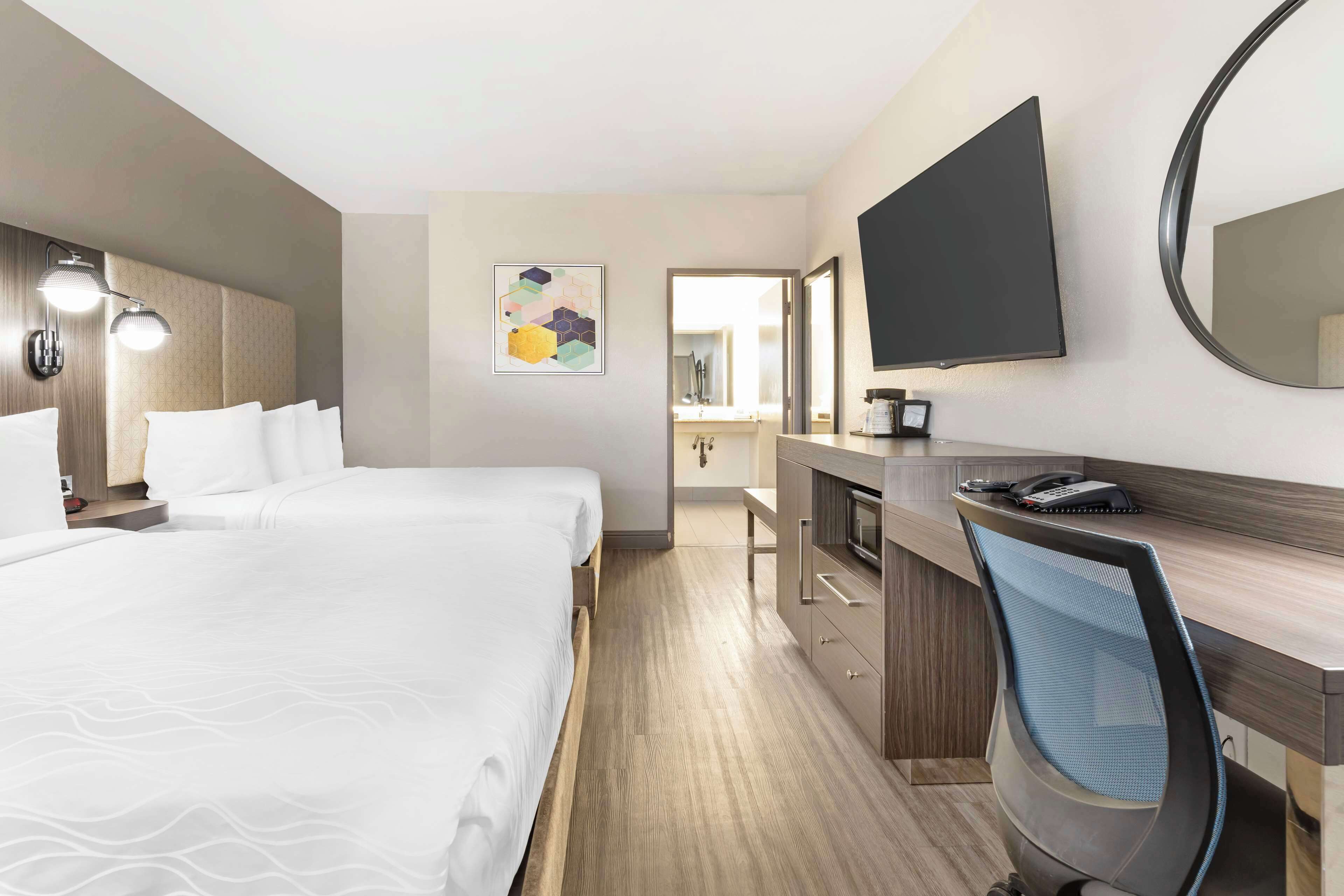 A hotel room with a large bed, a desk with a chair, a flat-screen TV, a mirror, and a bathroom visible through an open door.