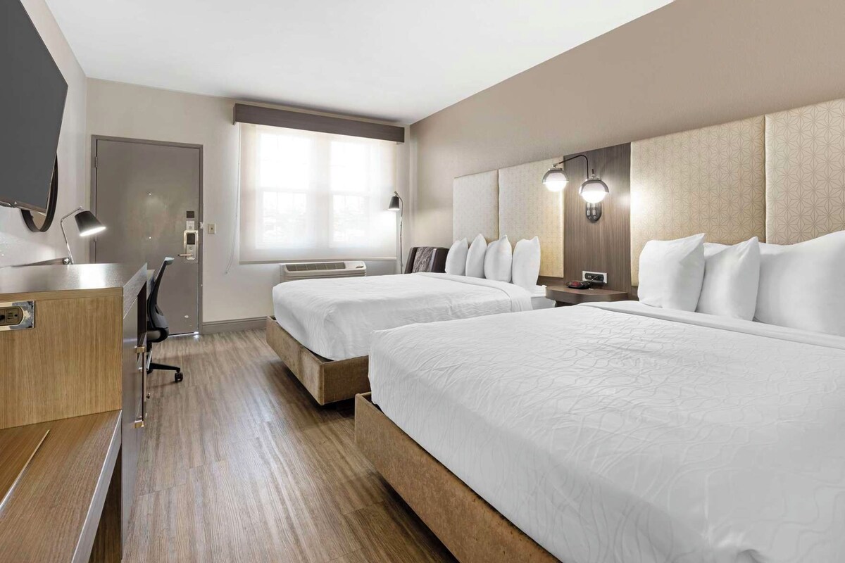 A hotel room with two beds, a desk, a chair, a window with blinds, and a wall-mounted lamp.