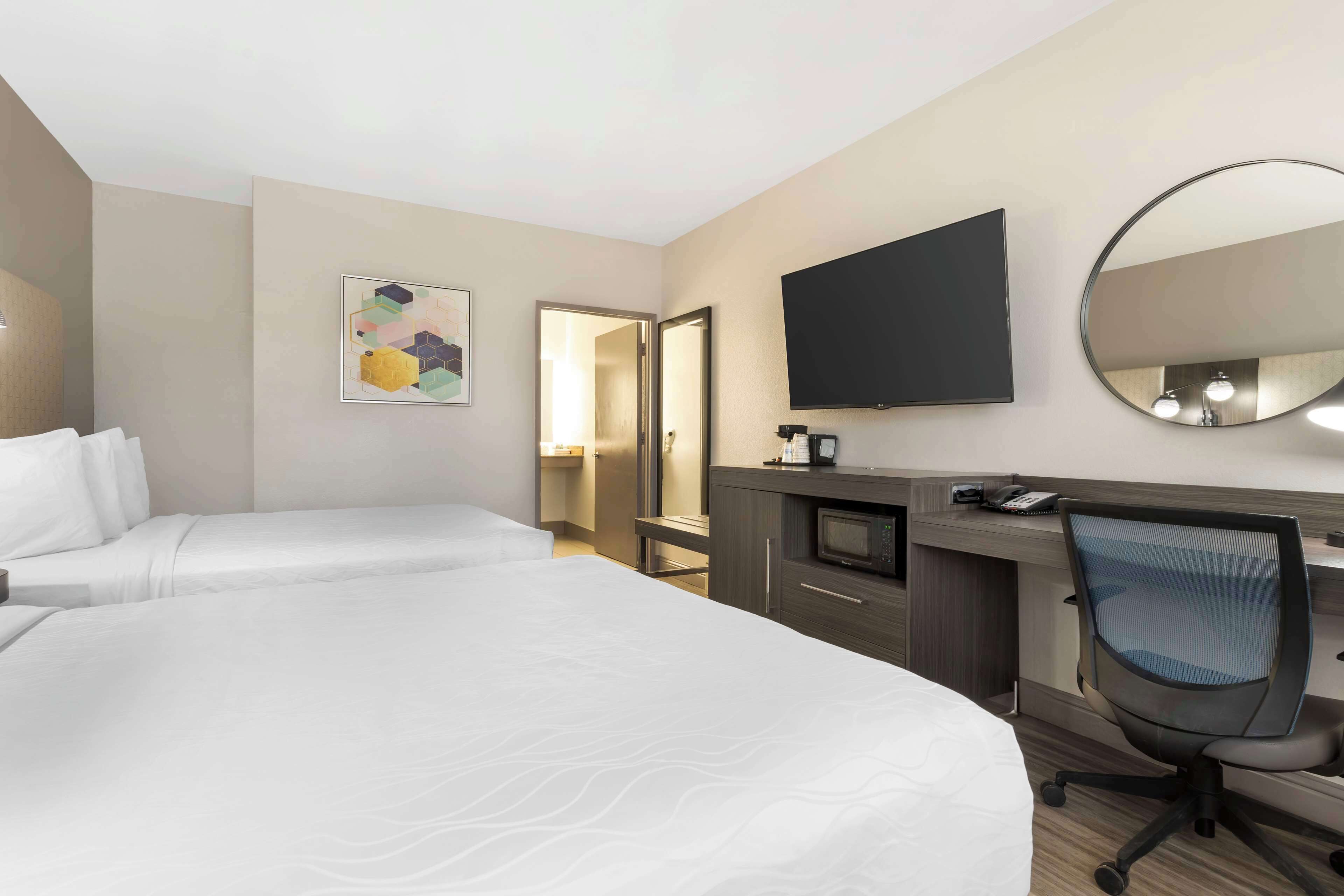 A hotel room with two beds, a desk with a chair, a flat-screen TV, a mirror, and a small kitchenette.