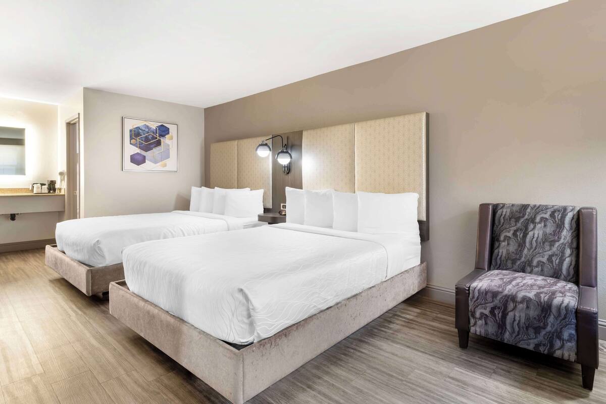 A hotel room with two beds, a headboard, a chair, and a wall-mounted lamp.
