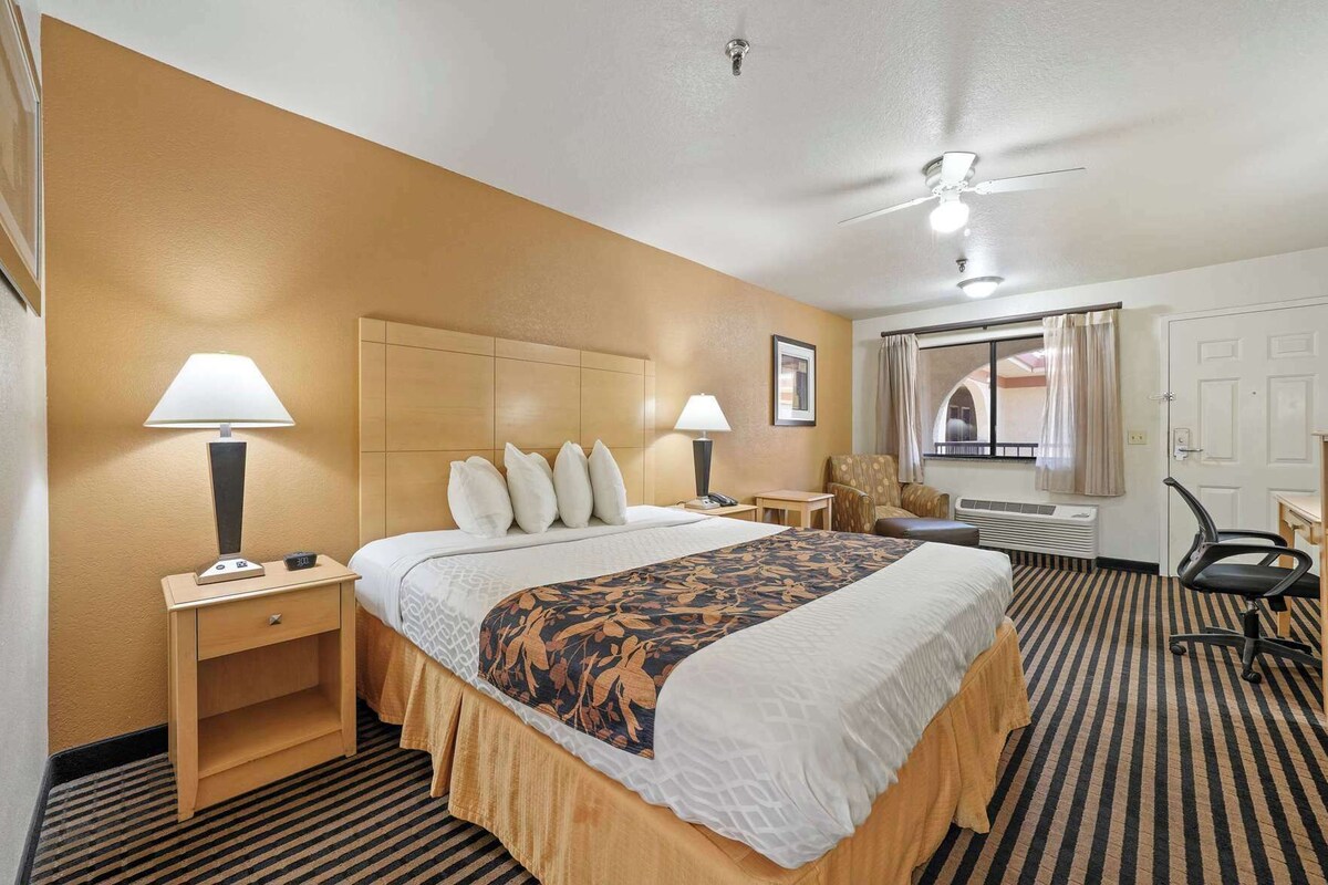 Habitación de hotel con una cama grande, dos mesitas de noche con lámparas, una silla, un escritorio, un ventilador de techo y una ventana con cortinas.