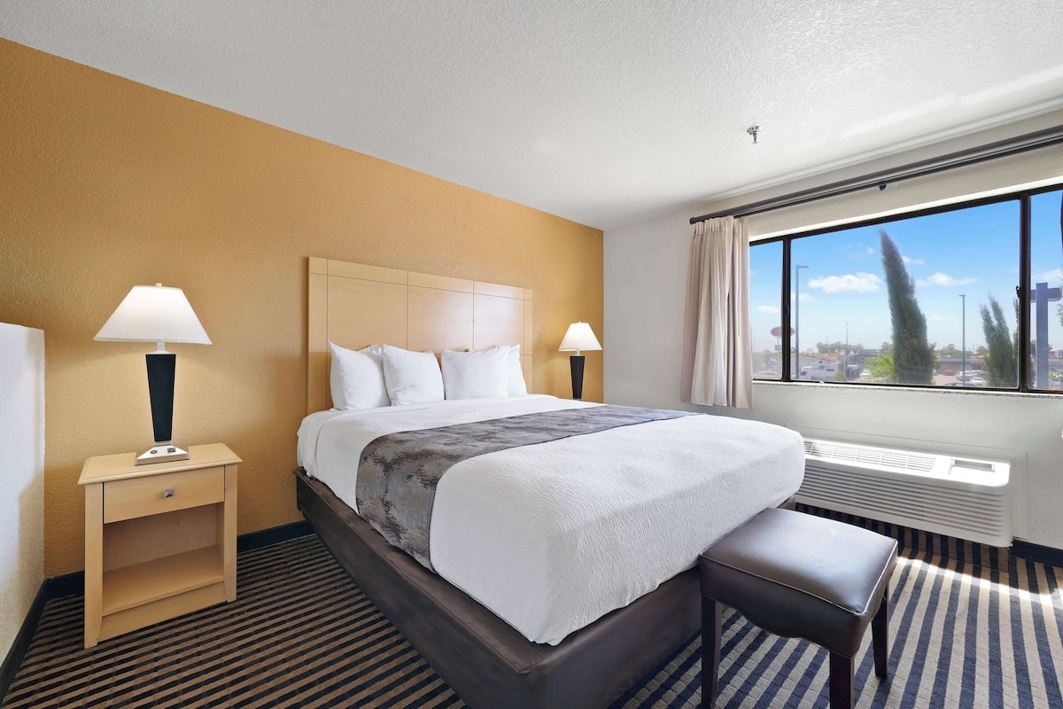 A hotel room with a large bed, two bedside tables with lamps, a bench, a window with a city view, and a wall-mounted air conditioner.