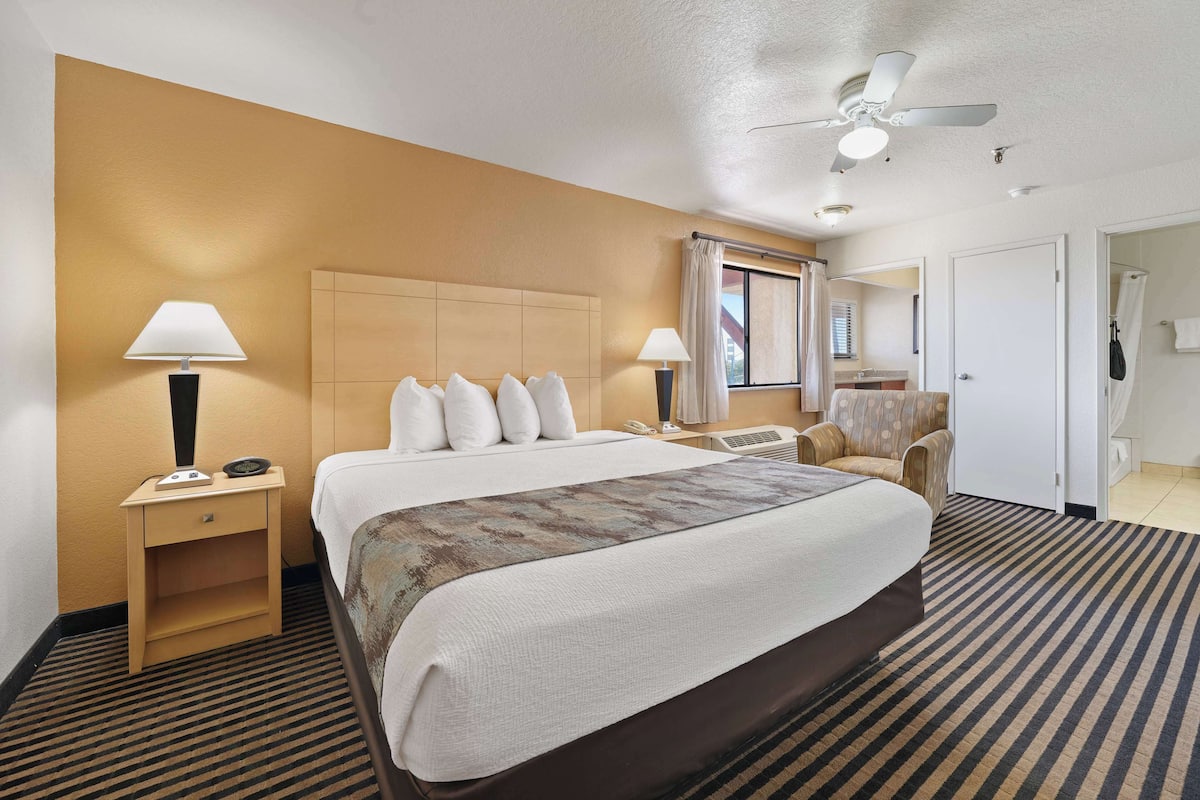 A hotel room with a large bed, two bedside tables with lamps, a ceiling fan, a window with curtains, and a bathroom visible through an open door.