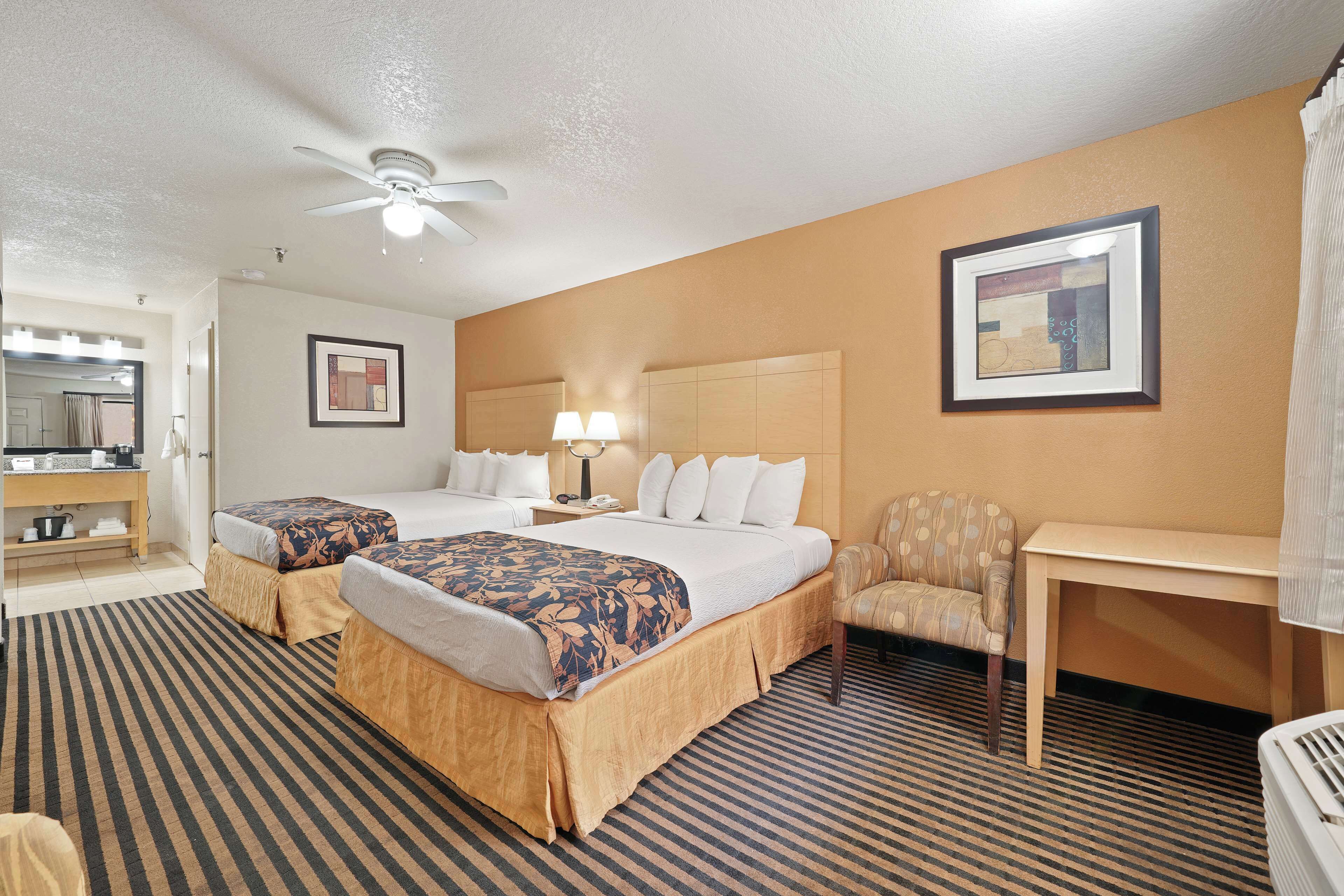 A hotel room with two beds, a desk, a chair, a ceiling fan, a framed picture on the wall, and a bathroom with a mirror and sink.