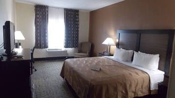 Habitación de hotel con una cama grande, dos lámparas de noche, una silla, un escritorio con computadora y televisión, y una ventana con cortinas.