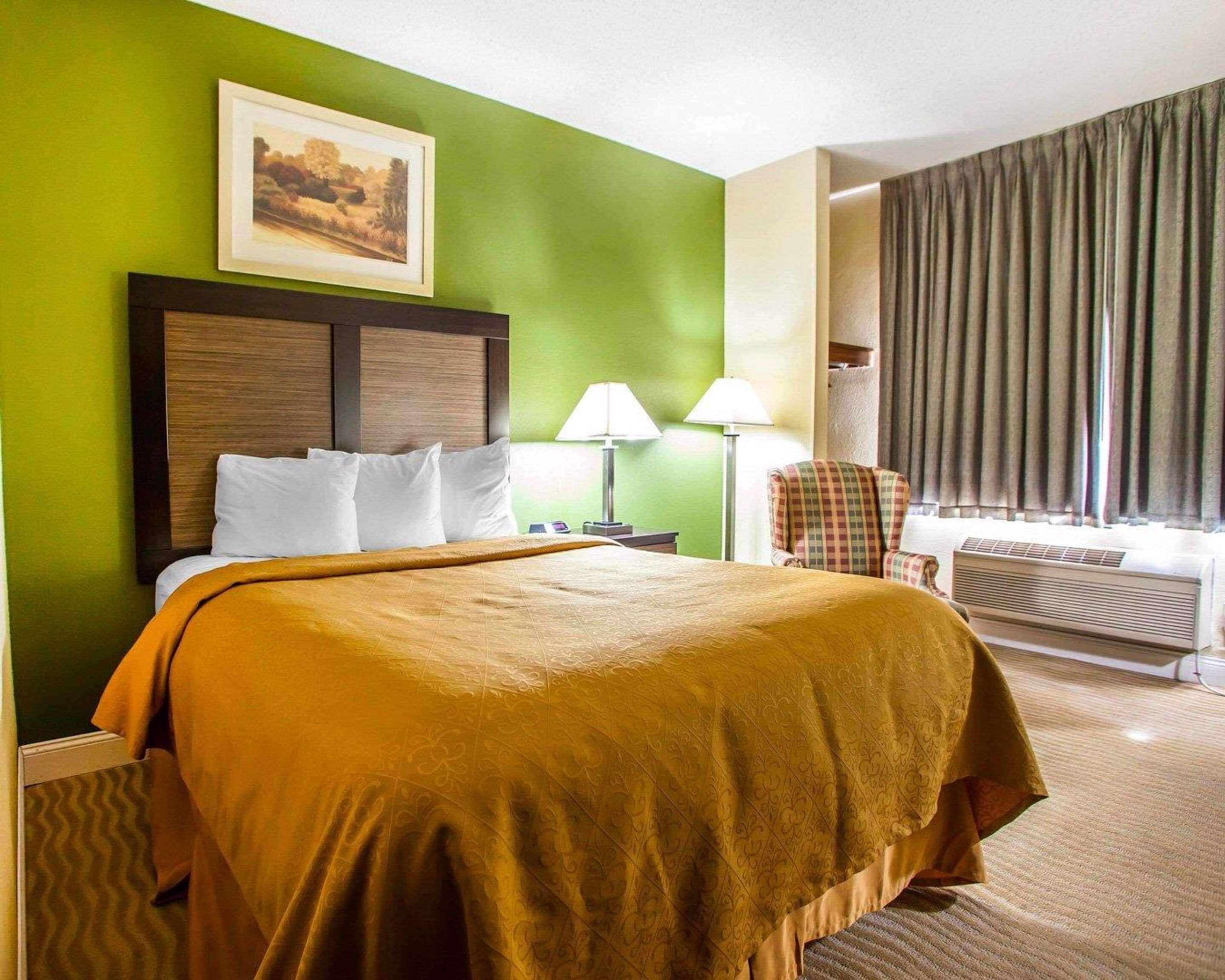 Habitación de hotel con cama, dos lámparas de noche, una silla, ventana con cortinas y un cuadro en la pared.