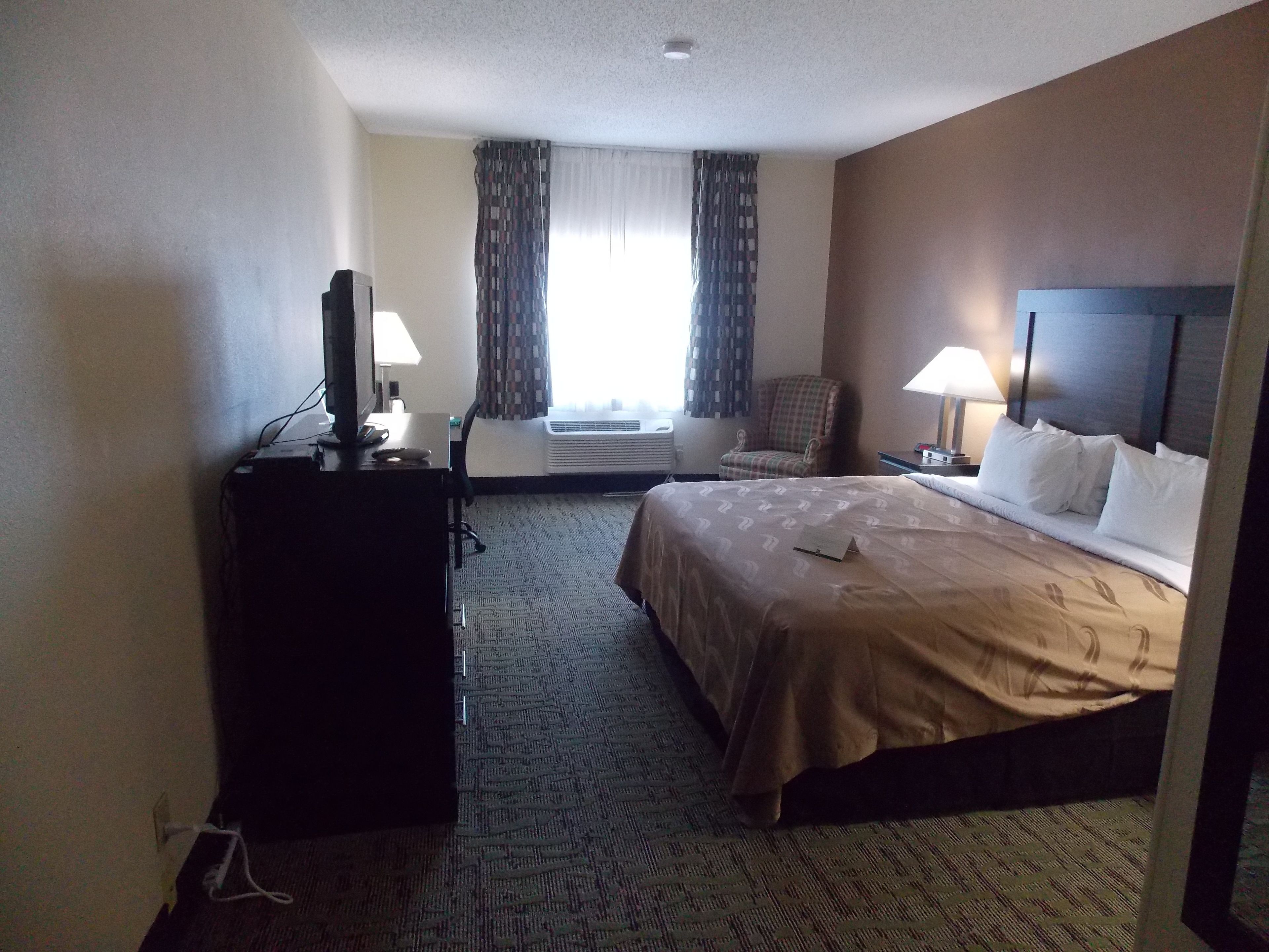Une chambre d’hôtel avec un lit, un bureau avec un ordinateur, une chaise et une fenêtre avec des rideaux.