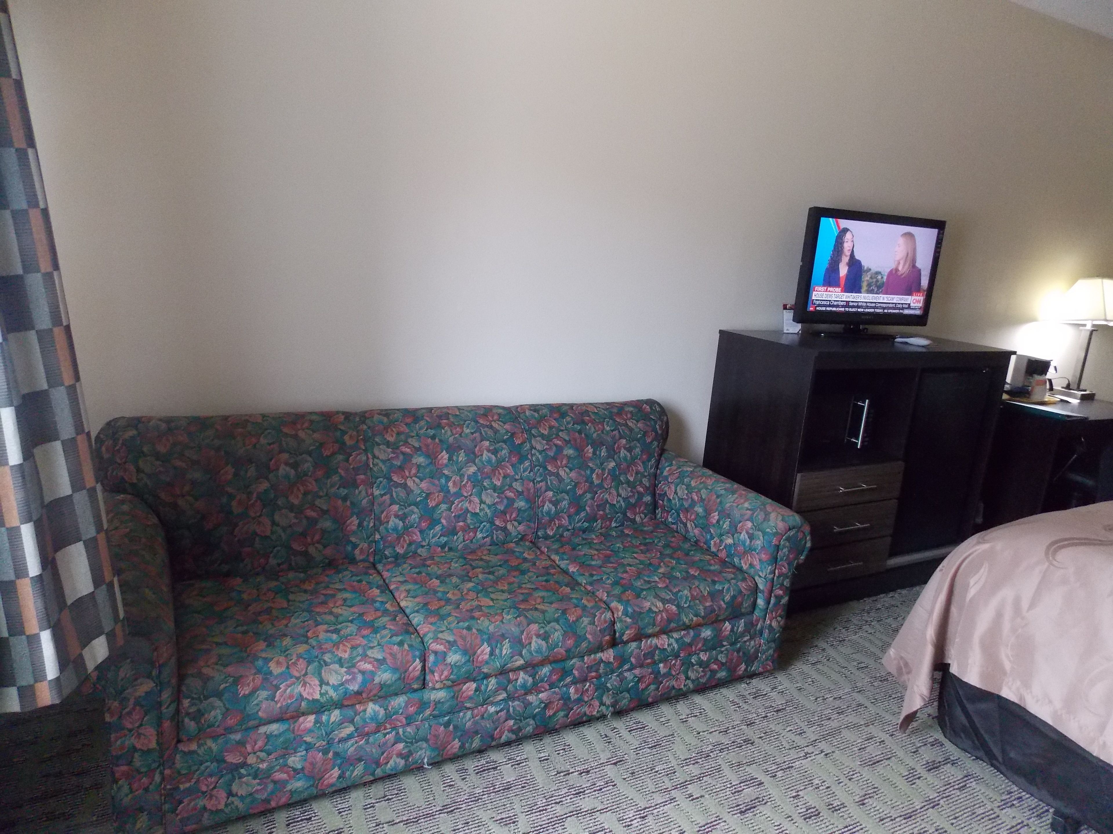 Une chambre d’hôtel avec un canapé à motifs floraux, un téléviseur posé sur un meuble et un lit recouvert d’une couvre-lit à motifs.
