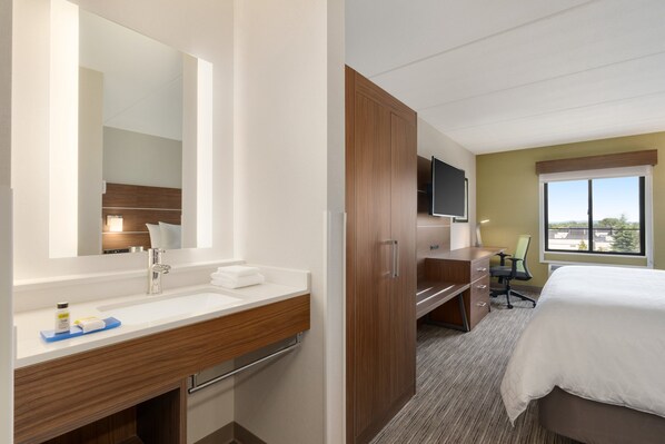 A hotel room with a large mirror, a wooden vanity, a bed, a desk with a chair, and a television.