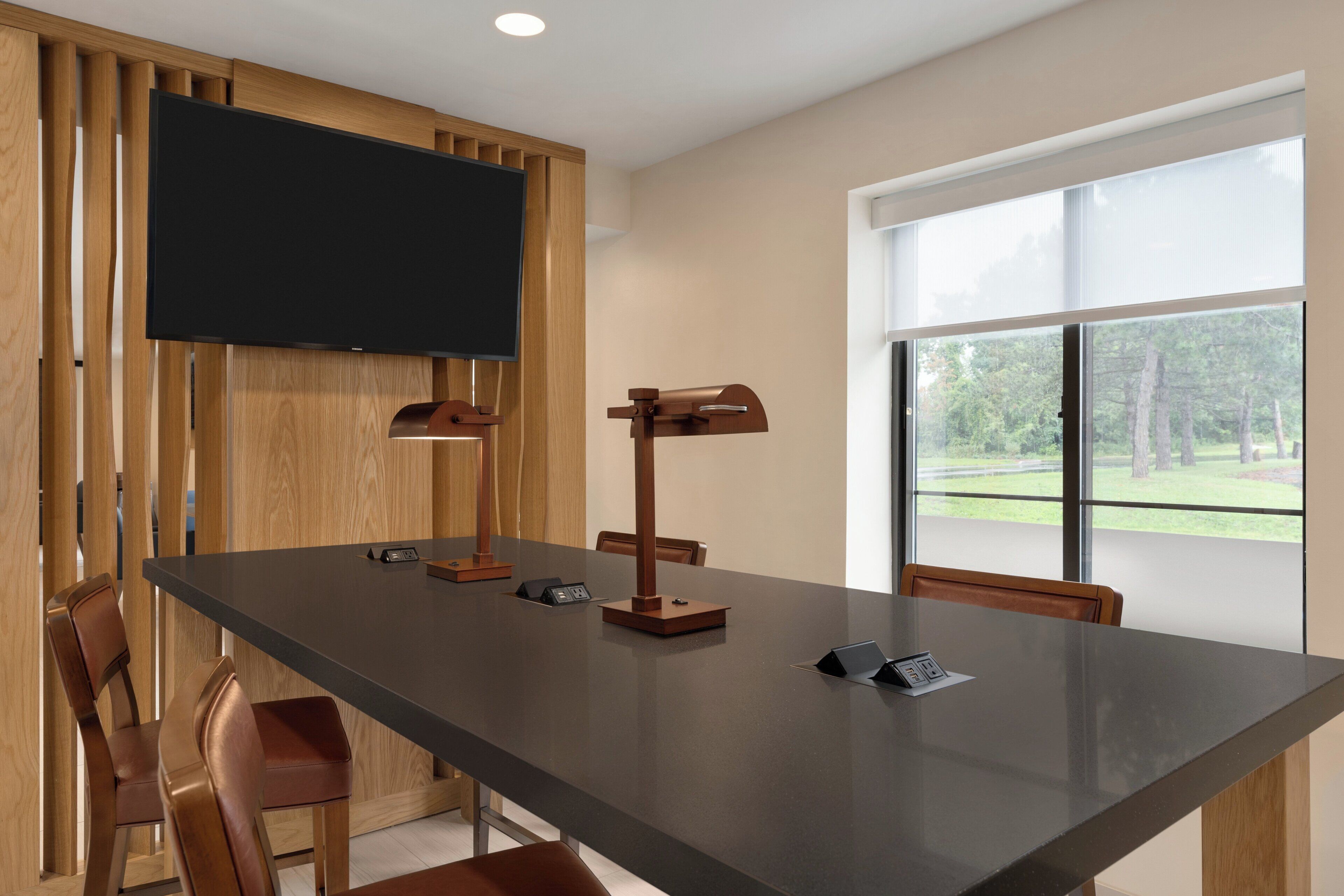 A conference room with a long table, chairs, a flat-screen TV, and a window with a view of trees.