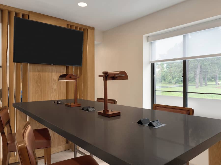 Un salón de conferencias con una larga mesa, sillas, un televisor de pantalla plana y un ventanal con vistas a árboles.
