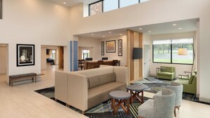A spacious living area with a high ceiling, a sofa, a coffee table, and a TV. There are framed artworks on the walls and a window providing a view of the outdoors.
