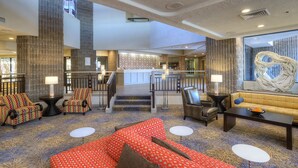 Eine großzügige Lobby mit hoher Decke, einer Treppe zu einer oberen Ebene und verschiedenen Sitzgelegenheiten, darunter Sofas und Sessel.