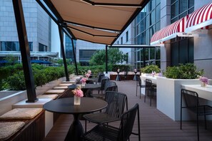 An outdoor patio with tables and chairs, a covered seating area, and a building with large windows in the background. - Kempinski Hotel Beijing Yansha Center (Beijing)