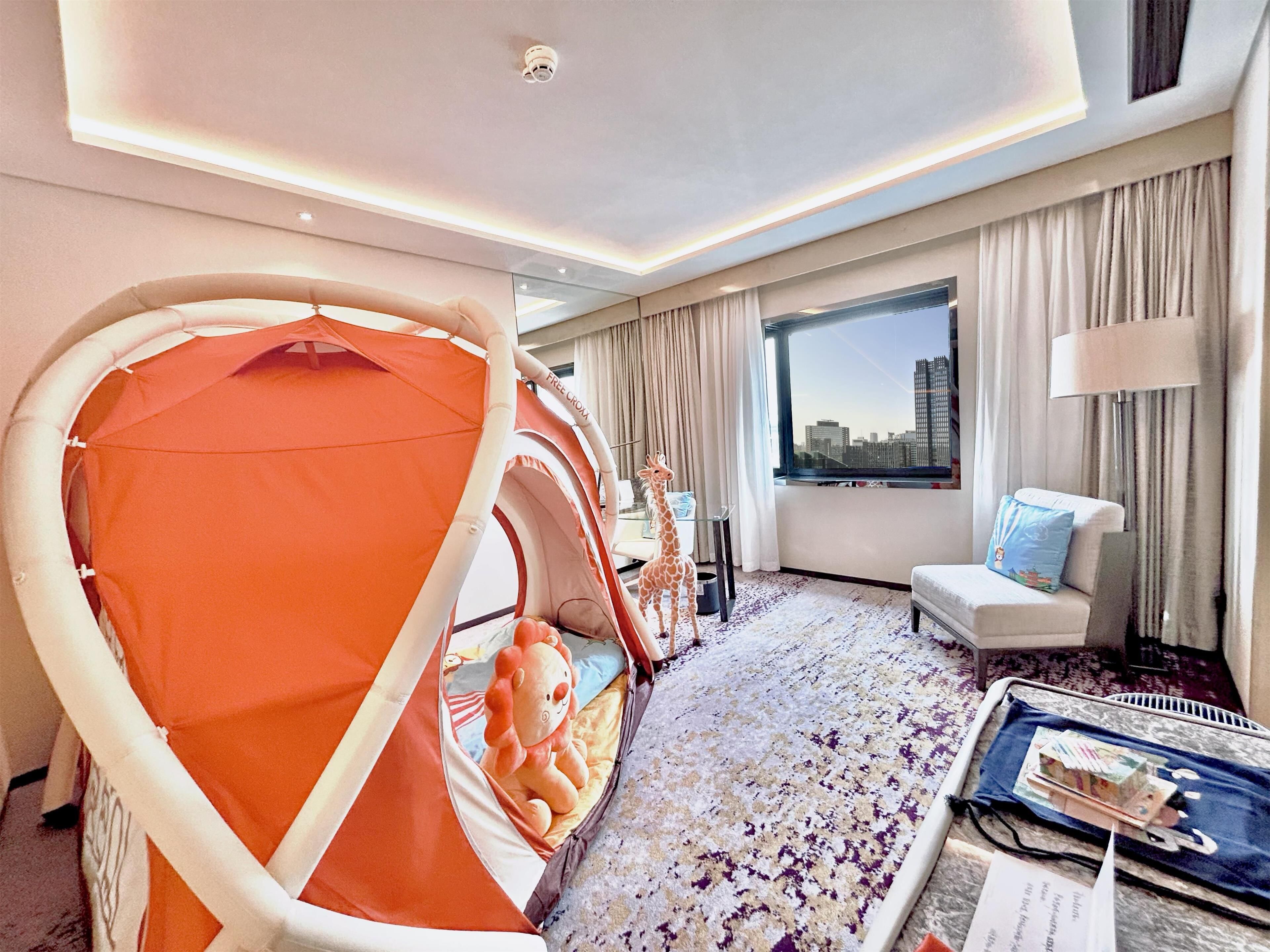 Ein Hotelzimmer mit großem Fenster, Blick auf die Stadt, einer Couch, einem Sessel, einem kleinen Tisch mit Gegenständen und einer zeltartigen Konstruktion mit einem Teddybären darin.