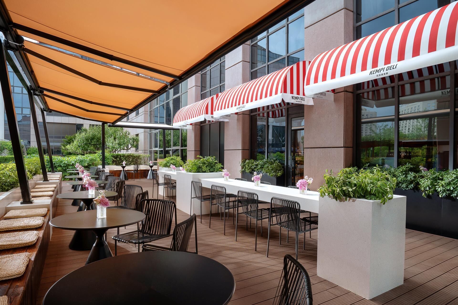 An outdoor seating area with tables and chairs, a covered section with a striped awning, and potted plants.