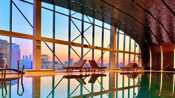 A modern indoor swimming pool with clear glass walls, lounge chairs, and a city view. - Kempinski Hotel Beijing Yansha Center (Beijing)