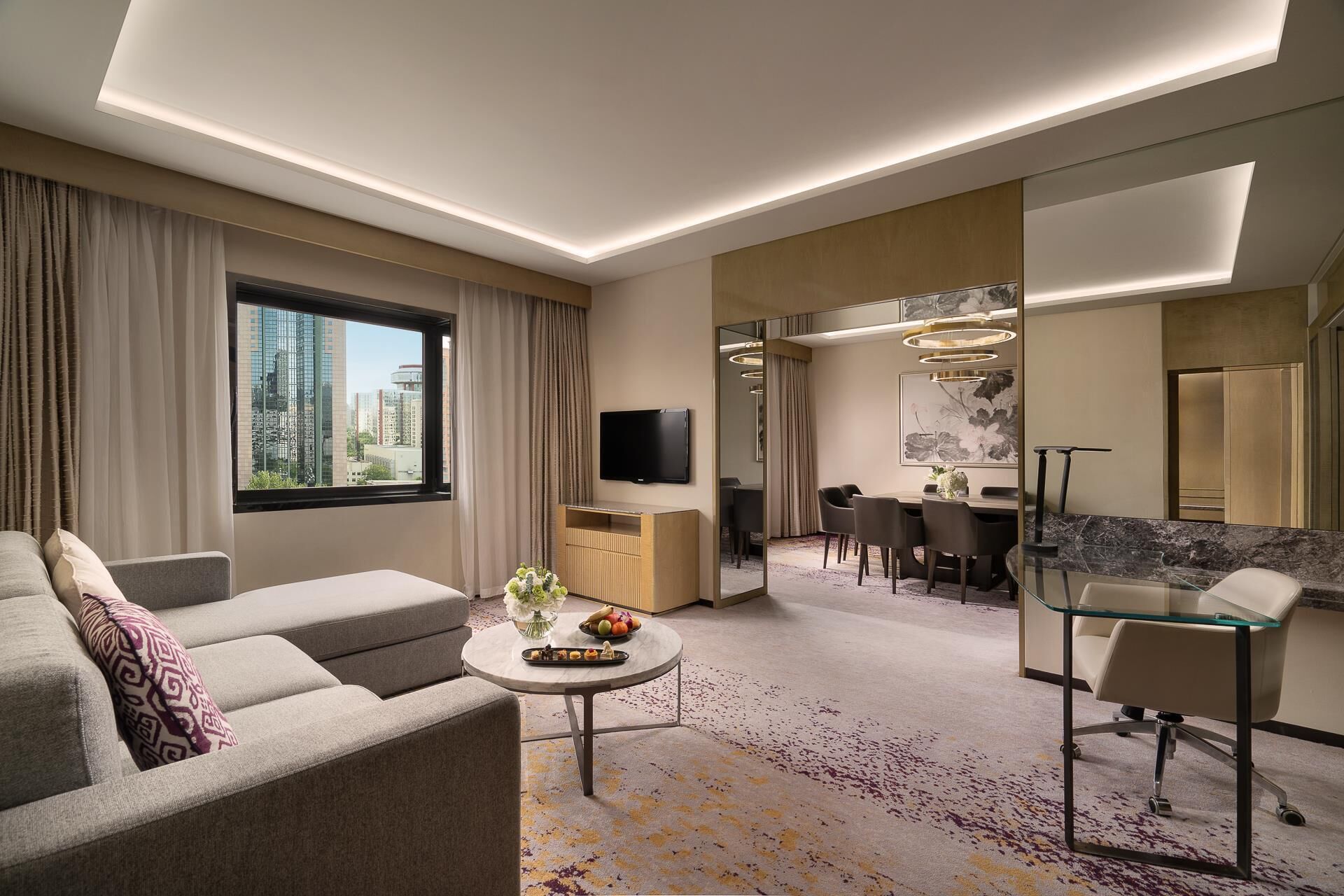 Una habitación de hotel moderna con sofá, una mesita redonda con un florero y fruta, un área de comedor con mesa y sillas, y un espejo grande.