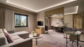 Una habitación de hotel moderna con sofá, una mesita redonda con un florero y fruta, un área de comedor con mesa y sillas, y un espejo grande.
