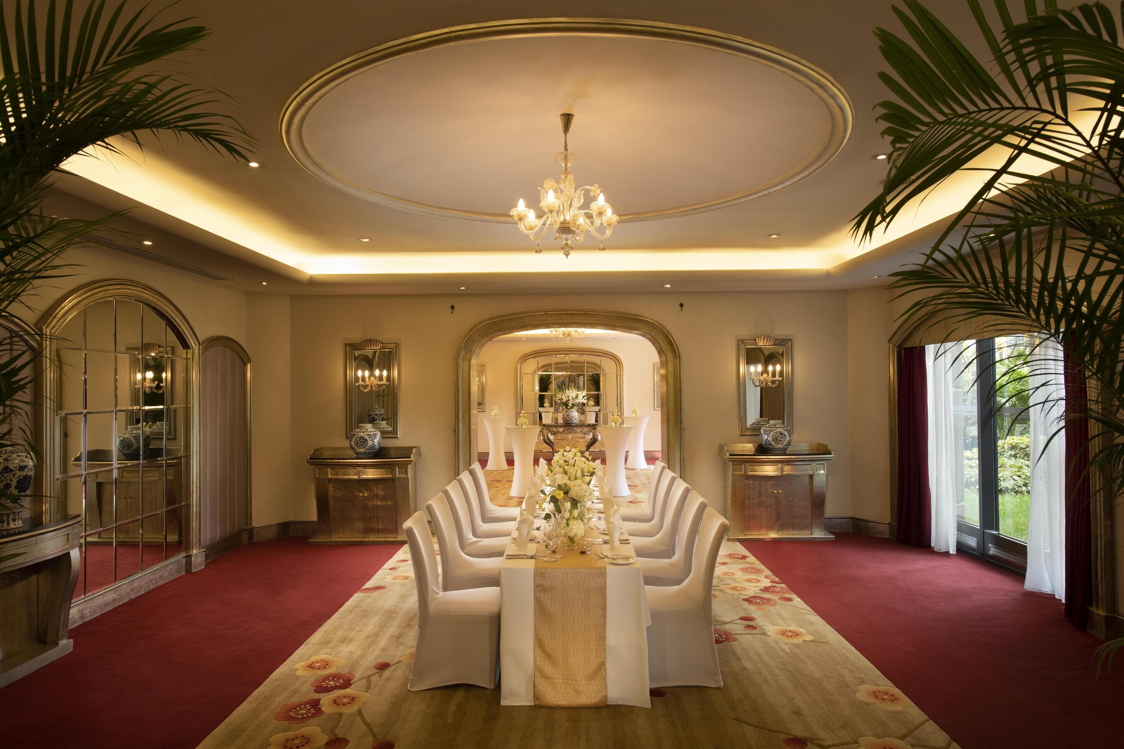 A formal dining room with a long table set for multiple guests, elegant chandeliers, and large windows with curtains.