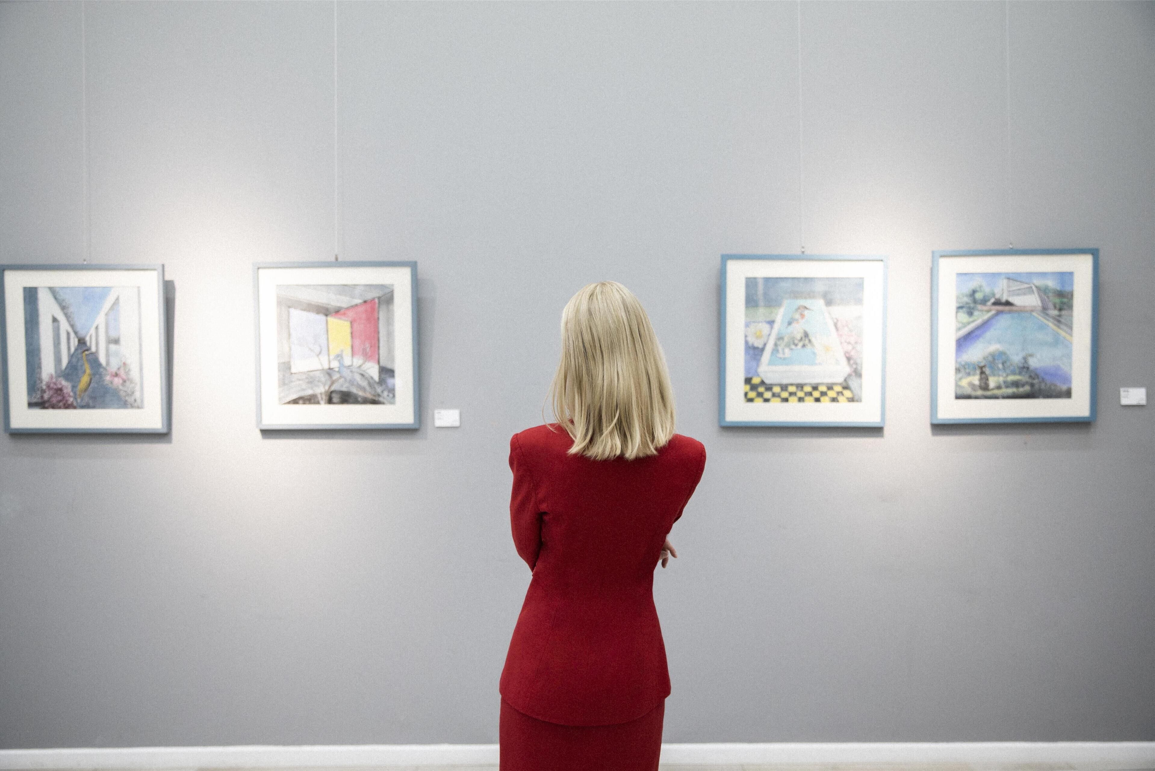 Eine Person in einem roten Anzug betrachtet gerahmte Kunstwerke an einer Ausstellungswand.