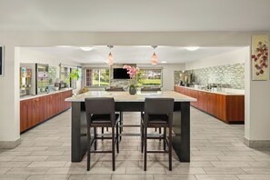 Una cocina moderna con una isla central, dos taburetes de bar, armarios de madera, un ventanal grande y un televisor de pantalla plana.