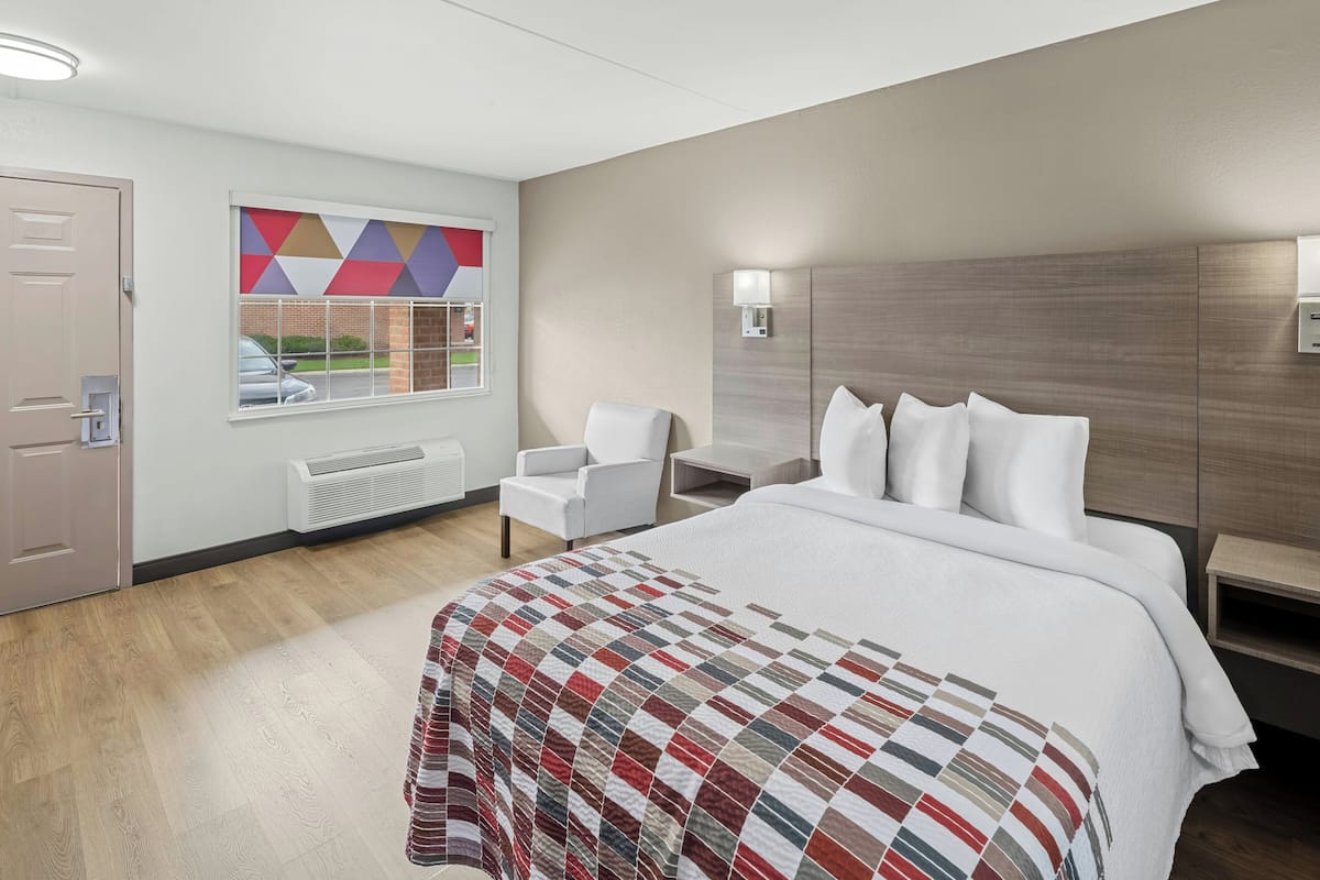 Une chambre d’hôtel comprenant un lit, une chaise, un bureau, une fenêtre donnant sur un paysage aux formes géométriques et un luminaire fixé au mur.