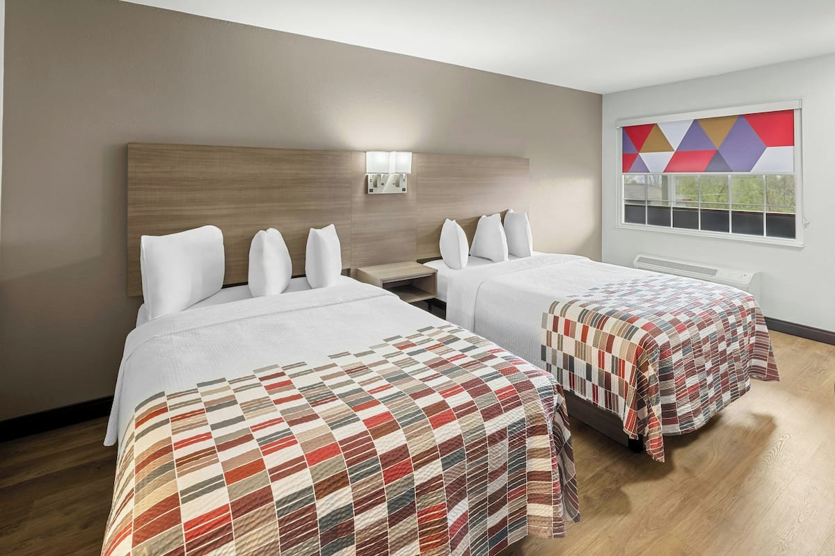 Une chambre d’hôtel avec deux lits, une table de chevet, une lampe fixée au mur et une fenêtre donnant sur un paysage aux formes géométriques.