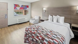 A hotel room with a bed, a chair, a desk, a window with a geometric view, and a wall-mounted light.